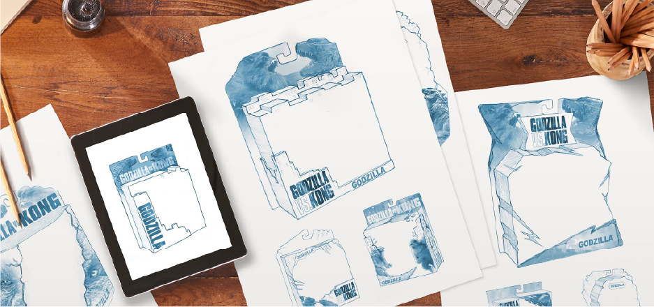 Several blue pencil sketches of Godzilla vs Kong toy packaging concepts are spread out on a wooden desk. The sketches show different box shapes and layouts, each labeled with Godzilla vs Kong and Godzilla. An electronic tablet displays one of the sketches. The scene feels creative and energetic, suggesting a design brainstorming session.